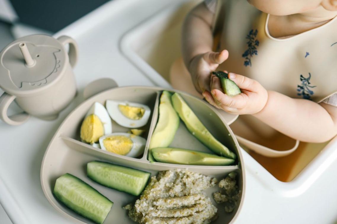 Baby sitting in a high chair, eating foods including egg, avocado, cucumber.